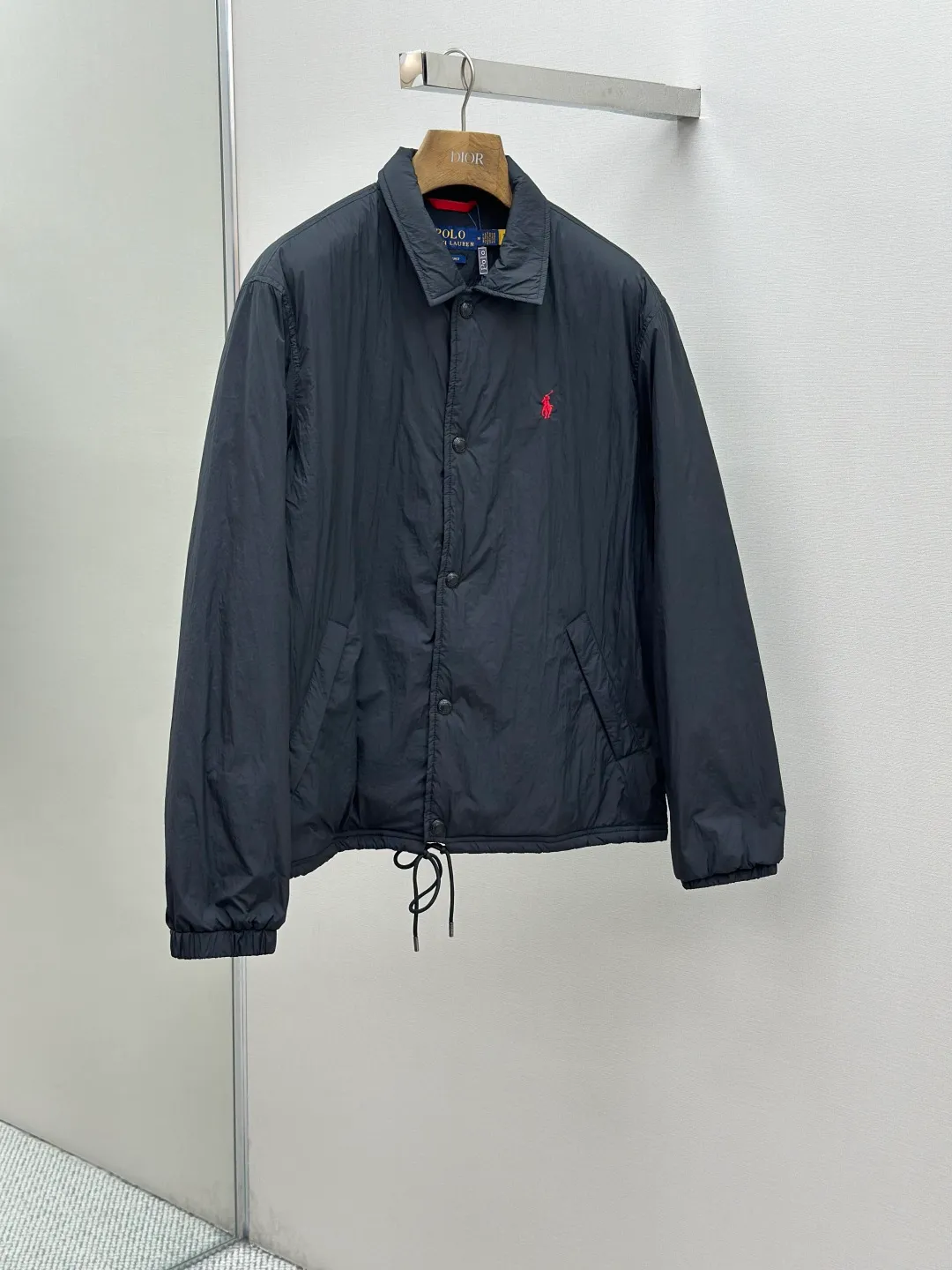 P650 RAH LAUREN RL red label lapel is light and thin. It is made of high-density nylon taffeta with a density of about 210T-300T. The surface is smooth and slightly matte. It has excellent windproof and waterproof performance (with R water-repellent treatment). It has strong wrinkle resistance and is easy to care for. It adopts the vat dyeing process (the dye penetrates into the fiber) and the color fastness reaches ISO level 4 or above to avoid fading or color transfer after washing. The lightweight cotton-polyester blend lining (65% Polyester + 35% Cotton) reduces the friction between the fabric and inner clothing and improves the smoothness of wearing. The blended material combines breathability and durability to avoid the stuffy feeling of pure nylon lining. The classic coach (Jaet) loose fit has a slightly dropped shoulder line (the shoulder width is 1-2cm wider than the standard fit), the length reaches the hips, and the sleeves are slightly tapered straight sleeves, taking into account freedom of movement and casual style. Flat satin stitch embroidery (satin embroidery), using 100% polyester high color fastness embroidery thread (the color number is consistent with the brand's standard red color card), the stitch density is 12-15 stitches cm, the outline of the horse logo is clear, and the details of the rider and horse are burr-free. Precise positioning through the template (12cm from the bottom of the collar and 8cm from the placket) ensures symmetry and centering. The seams of the body are made of double-needle flat seams (needle pitch 3cm, 10 needles), the stitches are flat and straight, the stitching color is dark black (the same color as the fabric), and the stitch width is 0.5cm, which enhances firmness and forms simple decorative lines. The placket is made of double-layer folding + flat seam splicing, with a thickness of about 0.3cm to ensure that the buttons are not easily deformed when fixed; the edges of the placket are cut with a hot knife to avoid burrs. The armholes are machine stitched with round head keyholes and flat seams. After stitching, the armholes have a natural curvature and no sense of tightness during movement. The height of the sleeves is about 8-10cm, which is in line with the casual and loose characteristics. Black urea resin button with matte surface treatment. The buttonholes are hand-made keyholes (6 strands of cotton thread, keyhole density of 8 stitches mm), and the edges of the eyelets are smooth and without burrs. The first button on the neckline is a hidden button (diameter 12mm), which prevents the neckline from opening and improves the neatness of wearing. Size:S-XL